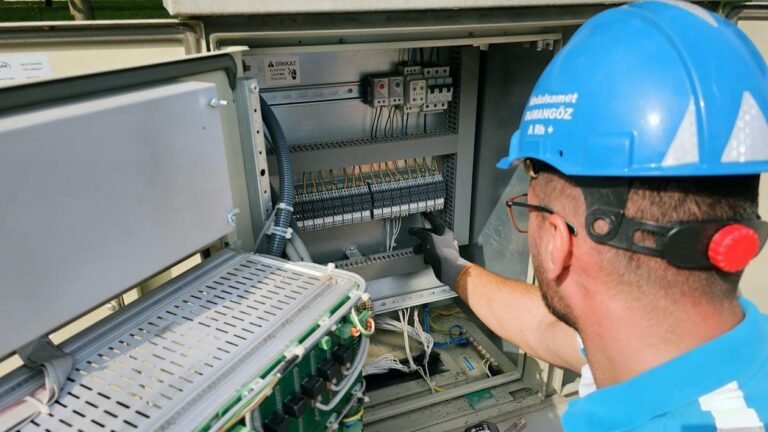 Technician wearing a blue hard hat, inspecting and adjusting breakers inside an open commercial electrical panel